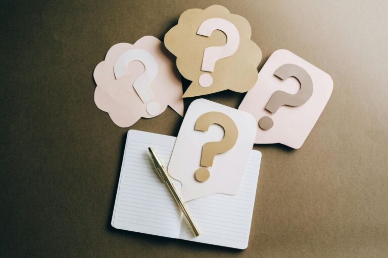 Cardboard question marks are positioned above an open notebook and pen.